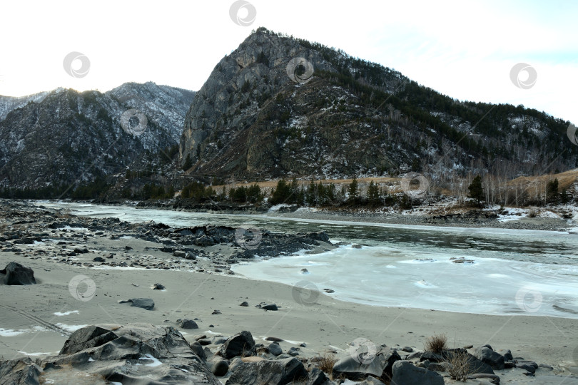 Скачать Каменистый берег с песком красивой замерзшей реки, текущей зимним вечером у подножия высокой скалы. фотосток Ozero