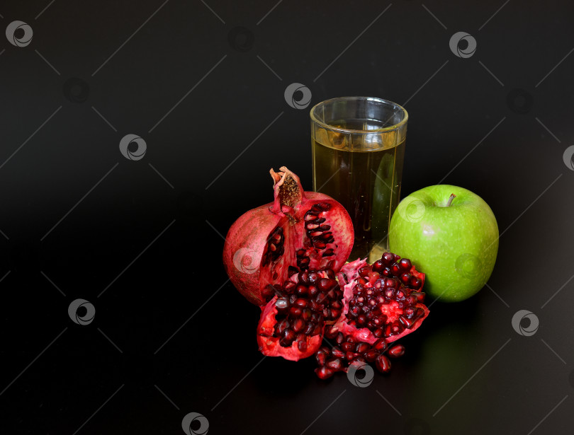 Скачать Высокий стакан со смешанными фруктовыми соками на черном фоне, спелое зеленое яблоко и разломанный плод граната с косточками. фотосток Ozero
