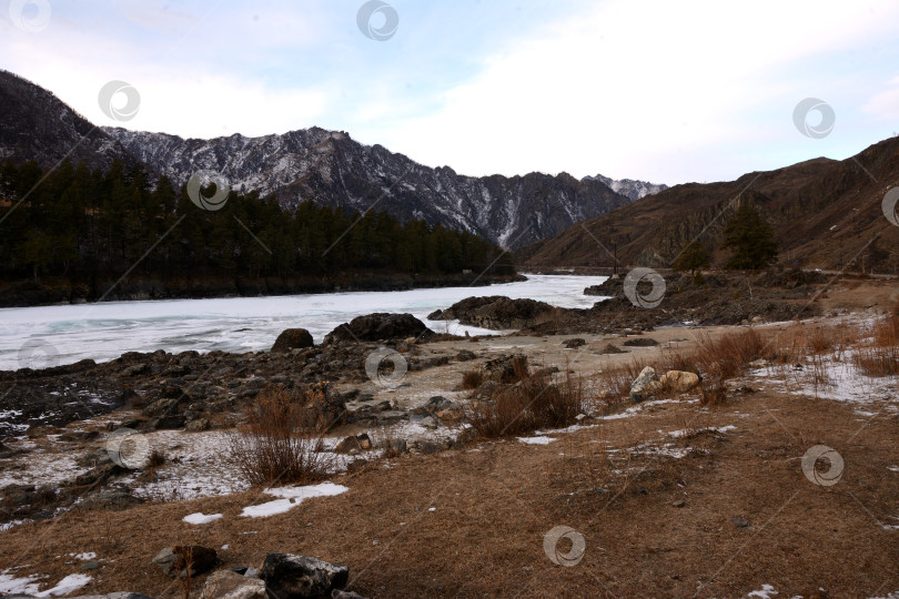 Скачать Скалистый берег с низкими кустами замерзшей реки, окруженный заснеженными горами, пасмурным зимним вечером. фотосток Ozero