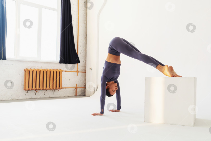 Скачать Женщина в спортивной одежде выполняет стойку на руках и опирается ногами на куб, тренируется в световой студии фотосток Ozero