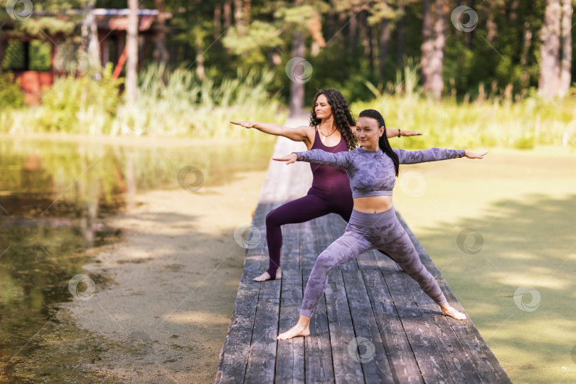 Скачать Две женщины, практикующие йогу, выполняют упражнение вирабхадрасана, поза воина, тренируются, стоя на деревянном мосту в парке фотосток Ozero