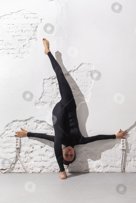 Скачать Женщина в черном спортивном комбинезоне выполняет упражнения на растяжку мышц ног, делая продольный шпагат, прислонившись спиной к стене в комнате фотосток Ozero