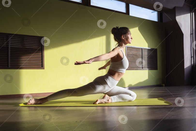 Скачать Молодая девушка, ведущая здоровый образ жизни, занимающаяся гимнастикой, тренирующаяся в белой спортивной одежде в комнате на коврике фотосток Ozero