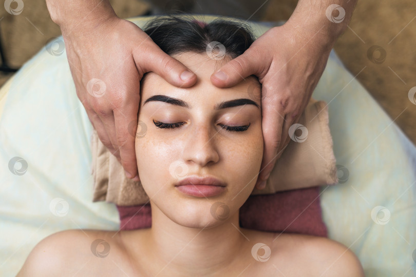 Скачать Специалист делает расслабляющий массаж лицевых мышц клиентке, крупным планом лицо сверху фотосток Ozero