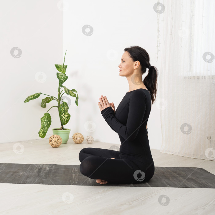 Скачать Женщина, ведущая здоровый образ жизни и практикующая йогу, медитирует, сидя в позе лотоса с намасте, тренируется в черной спортивной одежде, вид сбоку в полный рост фотосток Ozero