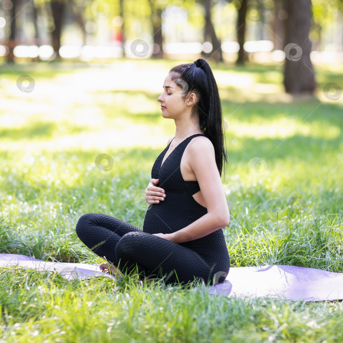 Скачать Молодая беременная девушка, ведущая здоровый образ жизни и практикующая йогу, медитирует, сидя в позе лотоса на коврике в парке солнечным теплым утром с закрытыми глазами фотосток Ozero