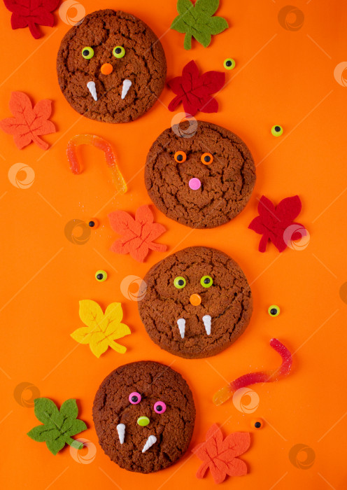 Скачать Печенье-монстр на Хэллоуин, украшенное сахарными глазами и клыками в окружении игрушечных осенних листьев на ярко-оранжевом фоне. Вид сверху. фотосток Ozero