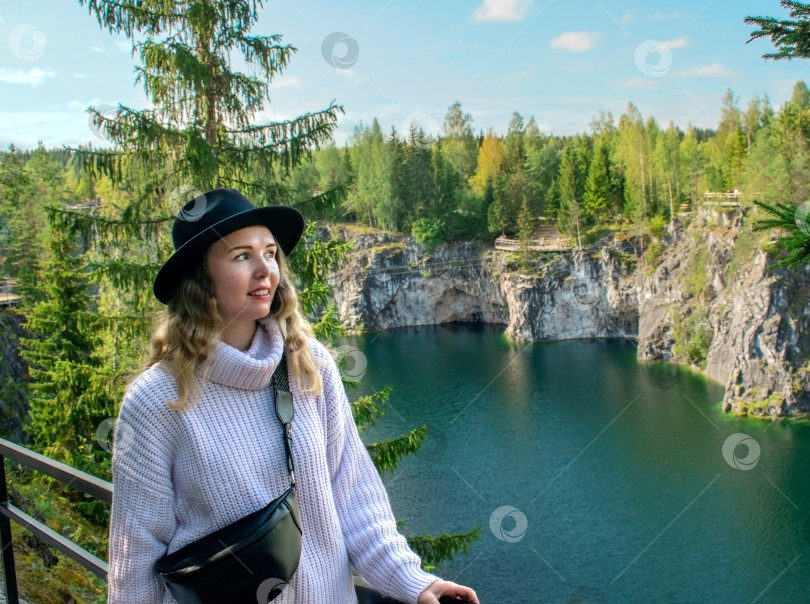 Скачать Молодая женщина в свитере, шляпе и поясной сумке улыбается, стоя у мраморного карьера с озером в парке Рускеала. фотосток Ozero