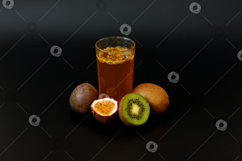 Скачать Высокий стакан свежевыжатого сока из тропических фруктов с косточками на черном фоне, рядом с кусочками киви и спелой маракуйи. фотосток Ozero