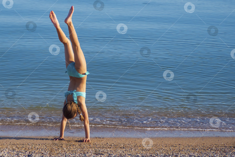 Скачать Ребенок на пляже занимается спортом и выполняет гимнастические упражнения на фоне моря. Девушка делает стойку на руках у кромки воды фотосток Ozero