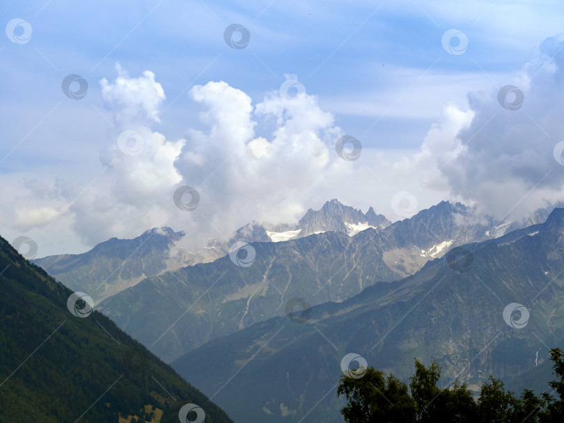 Скачать Горные хребты Альп расположены параллельно друг другу. Красивые облака лежат на вершинах гор. На ближайшем склоне растет хвойный лес. фотосток Ozero