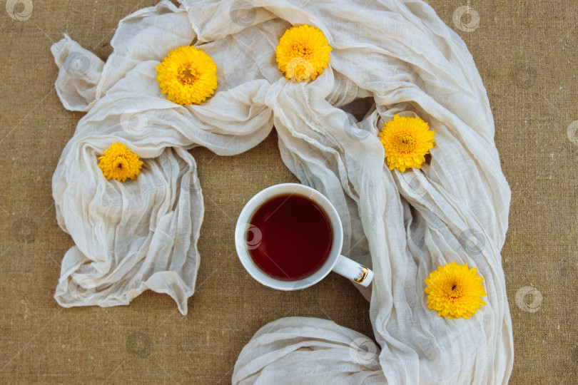 Скачать Белая кружка с чаем на столе и льняная скатерть с легким шарфом и желтыми цветами хризантемы вид сверху фотосток Ozero