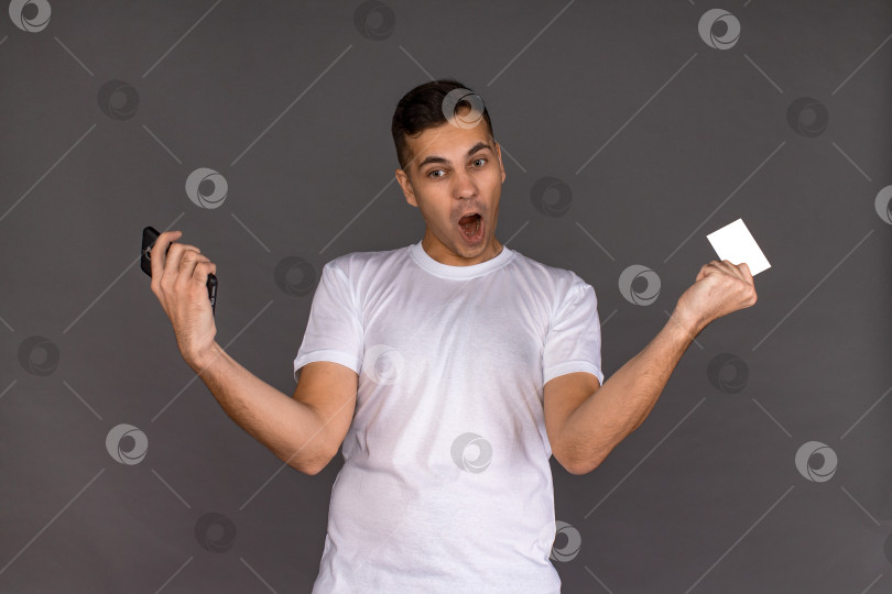 Скачать Молодой человек в белой футболке с телефоном и визитной карточкой счастлив фотосток Ozero