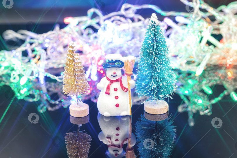 Скачать Снеговик стоит у рождественской елки на фоне сверкающей гирлянды. фотосток Ozero
