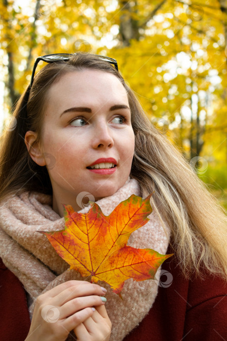 Скачать Молодая женщина в теплом шарфе и пальто держит в руках оранжевый кленовый лист и улыбается в парке. фотосток Ozero