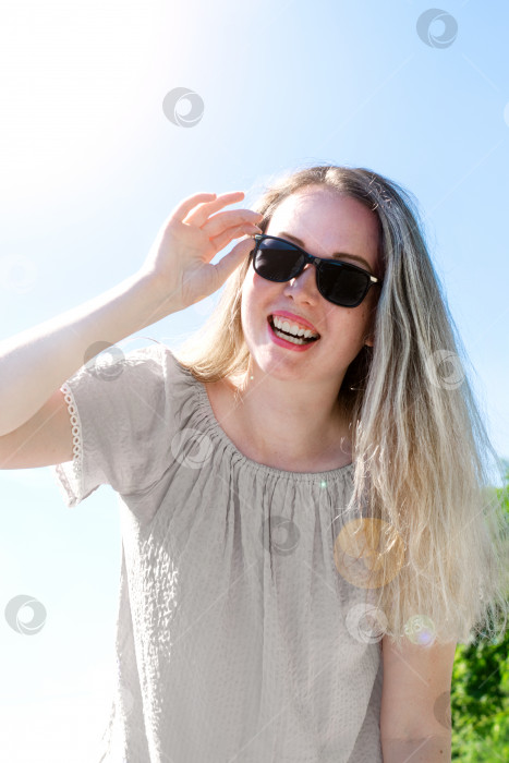 Скачать Молодая женщина со светлыми волосами с мелированием носит солнцезащитные очки, смотрит в камеру и улыбается летом. Солнце светит сбоку. фотосток Ozero