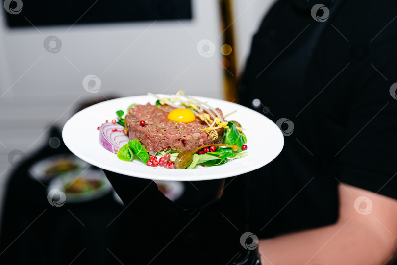 Скачать Официант держит тарелку с тартаром из говядины с салатом и сырым яичным желтком. Роскошная кухня, кейтеринг высокого класса. фотосток Ozero
