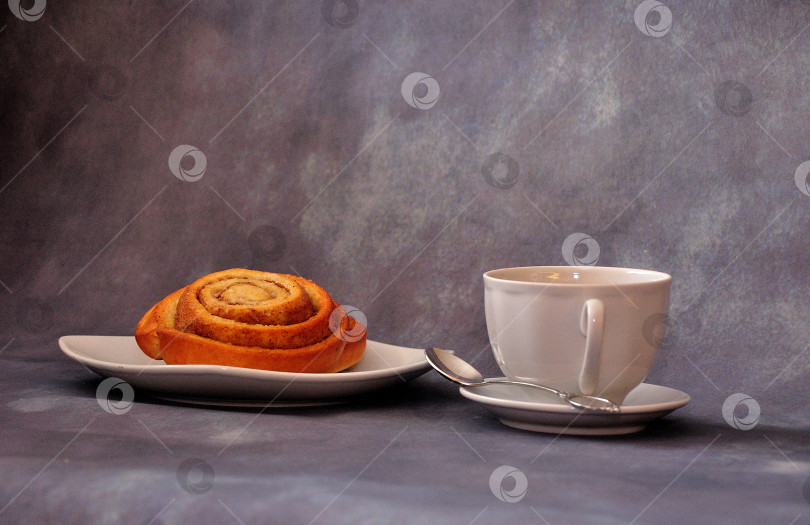 Скачать Скрученная булочка с сахаром и молотой корицей на тарелке и чашка горячего утреннего напитка на сером фоне. фотосток Ozero