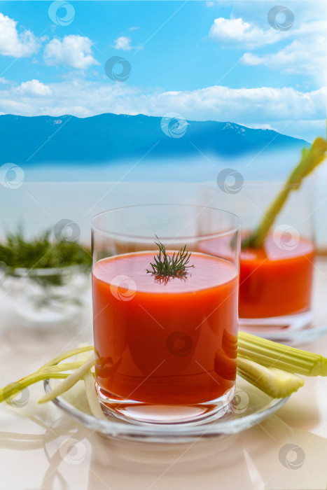 Скачать Подайте свежий томатный сок в стакане с палочками сельдерея и укропа на стол на открытом воздухе. Фон с горами и морем. фотосток Ozero