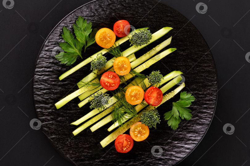 Скачать Салат из свежих овощей с огурцами, помидорами и зелеными листьями на черной тарелке на черном фоне. Нарезанные овощи. Вид сверху с местом для копирования. фотосток Ozero
