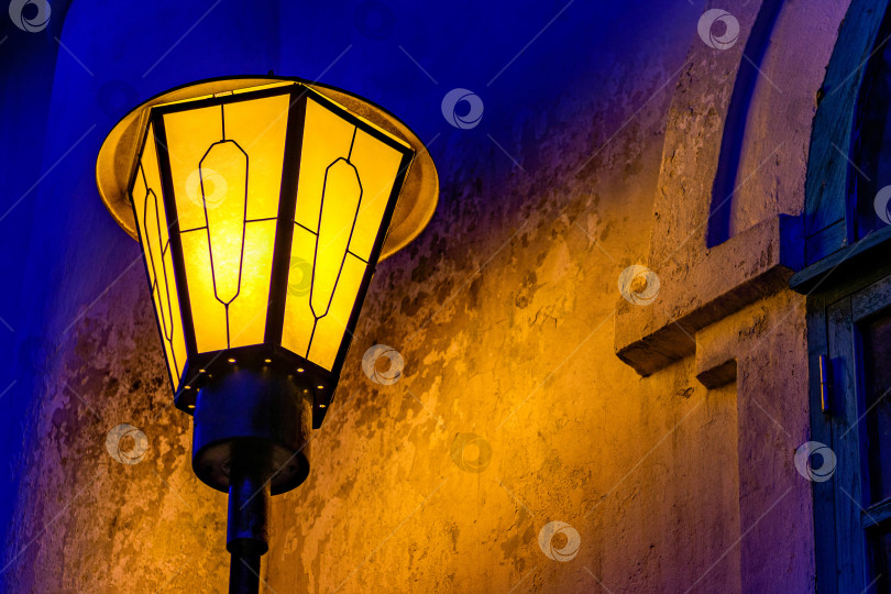 Скачать Старый винтажный уличный фонарь в селективном фокусе светится ночью. Волшебная лампа с теплым желтым светом в синих городских сумерках на фоне стены. Пространство для копирования. фотосток Ozero