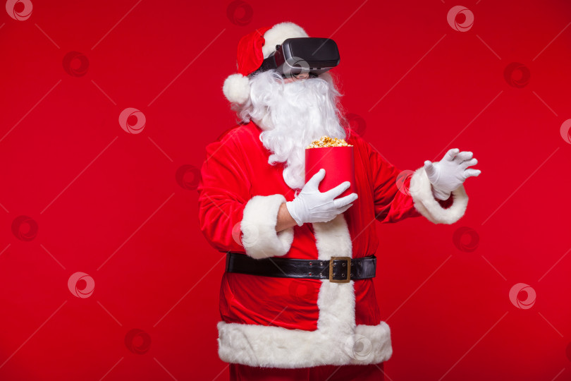 Скачать Санта-Клаус в очках виртуальной реальности и красном ведерке с попкорном на красном фоне. Рождество фотосток Ozero