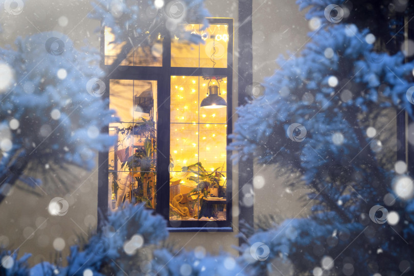 Скачать Уютное праздничное окно дома снаружи с теплым светом гирлянд сказочных огней внутри - празднуйте Рождество и Новый год в теплом доме. Рождественская елка, боке, снег на соснах и снегопад фотосток Ozero