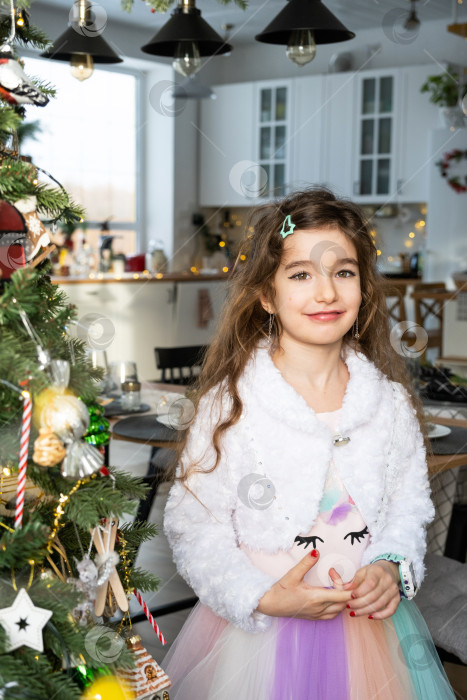 Скачать Портрет счастливой девочки-ребенка с длинными волосами в красивом платье в домашнем интерьере с рождественской елкой и новогодним декором. Подготовка к праздникам, вечеринкам фотосток Ozero