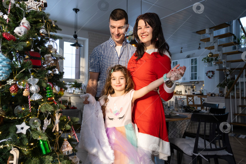 Скачать Традиционная семья из мамы, папы и дочери возле рождественской елки в интерьере своего дома. Празднуете Новый год со своей семьей фотосток Ozero