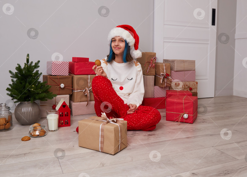 Скачать Счастливая девочка в пижаме и шапочке Санта-Клауса сидит у большой стопки рождественских подарков. фотосток Ozero