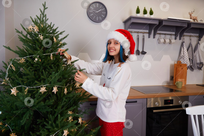Скачать Счастливая девочка в пижаме и шапочке Санта-Клауса украшает рождественскую елку к празднику. фотосток Ozero
