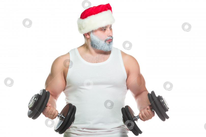 Скачать Крупный атлетически сложенный мужчина-спортсмен тренирует бицепсы, поднимая гантели, стоя в шляпе Санта-Клауса, готовый отпраздновать Рождество, изолированный на белом фоне фотосток Ozero