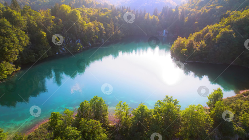 Скачать Горный лиственный лес со спокойной водой в озере на рассвете. Пейзаж с лета до ранней осени солнечным утром. фотосток Ozero