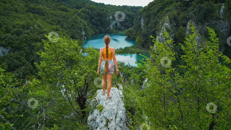 Скачать Спортсменка-скалолаз, девочка-подросток на вершине горы смотрит на озеро с бирюзовой водой и высокими водопадами. Туристка в Плитвицах, Хорватия. фотосток Ozero