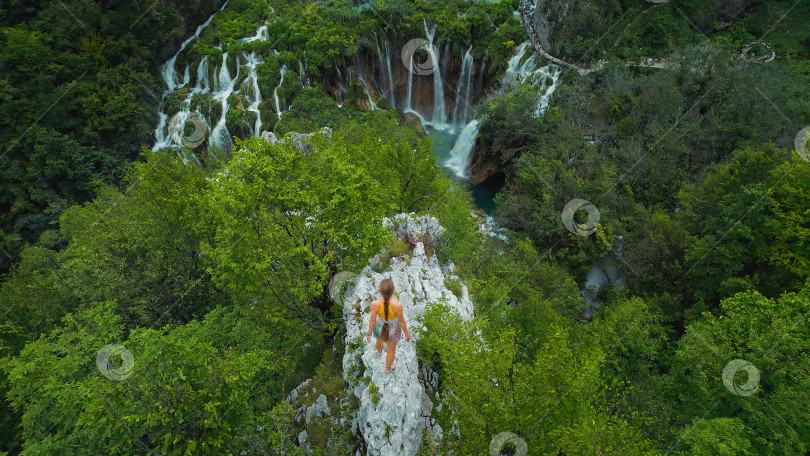 Скачать Спортсменка-скалолаз, девочка-подросток на вершине горы смотрит на озеро с бирюзовой водой и высокими водопадами. Туристка в Плитвицах, Хорватия. фотосток Ozero