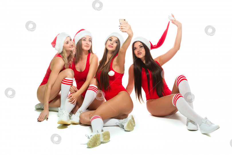 Скачать Счастливые молодые женщины миллениума весело празднуют Рождество, делая фотографии и селфи с помощью мобильного телефона, позируя, сидя на полу в красных купальниках и шляпах Санты, изолированных на белом фоне фотосток Ozero