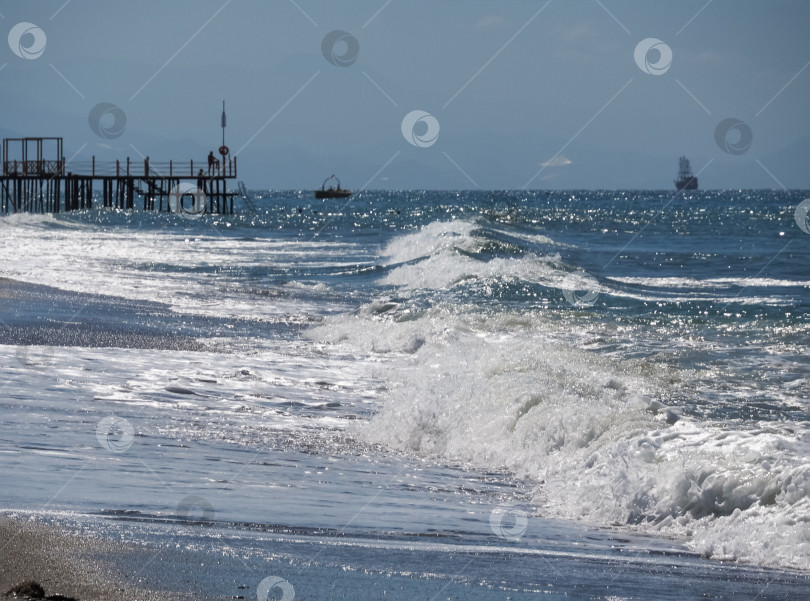 Скачать Морской пейзаж на заднем плане. Морские волны накатывают на берег. Вдалеке вы можете увидеть парусник, лодку и пирс. Сквозь дымку прорисовываются горы. фотосток Ozero