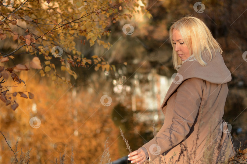 Скачать Счастливая женщина в желтом пальто с осенними листьями в руке и осенним желтым садом или парком. Красивая улыбающаяся молодая женщина в осенней листве. фотосток Ozero