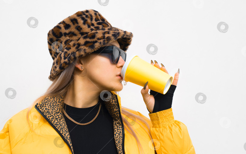 Скачать Молодая женщина пьет кофе из одноразового стаканчика на фоне белой стены снаружи, портрет в профиль. Модная девочка-подросток в желтой куртке и очках фотосток Ozero