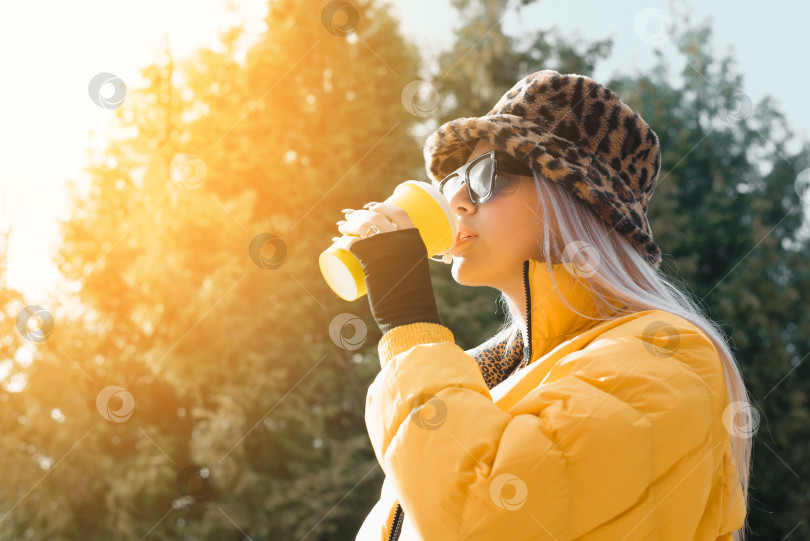 Скачать Молодая женщина пьет кофе из одноразового стаканчика на улице в солнечный день. Девочка-подросток в желтой куртке, очках и модной шляпке. фотосток Ozero