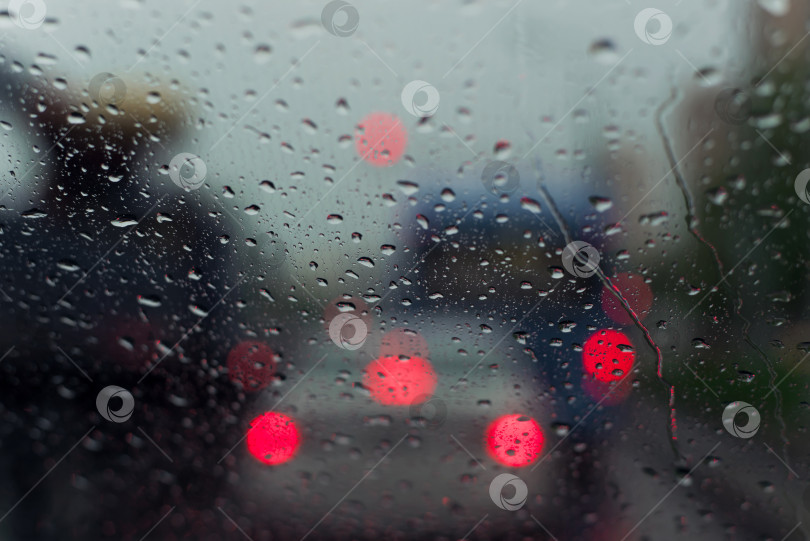 Скачать Капли дождя стучат по стеклу автомобиля. Размытые огни вечерней городской пробки. Мокрая поверхность лобового стекла автомобиля, капли стекают вниз, выборочная фокусировка. фотосток Ozero