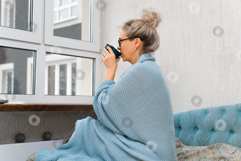 Скачать Женщина пьет кофе по утрам в помещении. Дама в очках сидит на кровати и закутана в теплое одеяло. Она держит кружку с горячим напитком и смотрит в окно. фотосток Ozero
