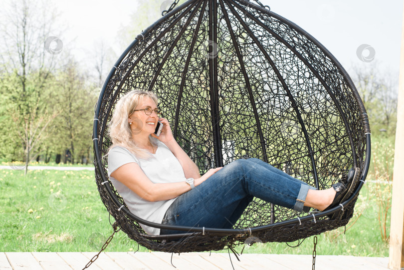 Скачать Счастливая женщина разговаривает по мобильному телефону и сидит на подвесных качелях в летнем парке. Улыбающаяся дама в очках отдыхает в городском саду. Отдых, концепция образа жизни. фотосток Ozero