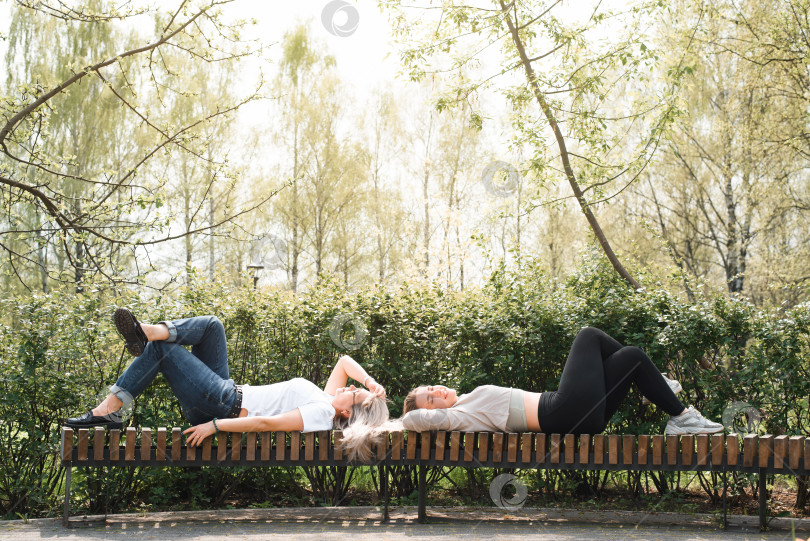 Скачать Две молодые женщины отдыхают на свежем воздухе. Подружки лежат на скамейке в городском саду. Счастливые девушки отдыхают в парке в солнечный день. фотосток Ozero