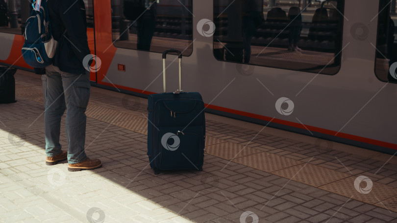 Скачать Вид сбоку на пассажира с рюкзаком и чемоданом, стоящего на платформе железнодорожного вокзала рядом с поездом в ожидании поездки, на открытом воздухе. Избирательное внимание к путешественнику, туристу фотосток Ozero