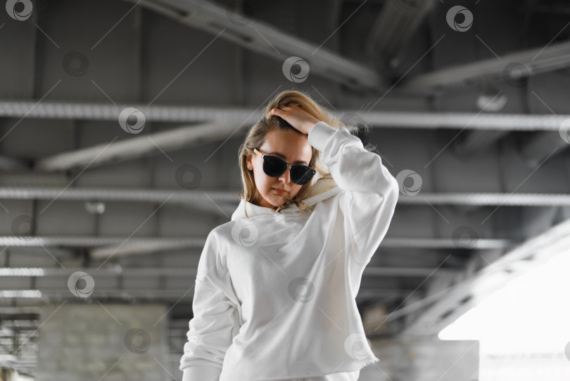 Скачать Стильный женский портрет в урбанистическом стиле. Женщина в солнцезащитных очках и светлой одежде позирует под мостом и поправляет волосы. Модная блондинка фотосток Ozero