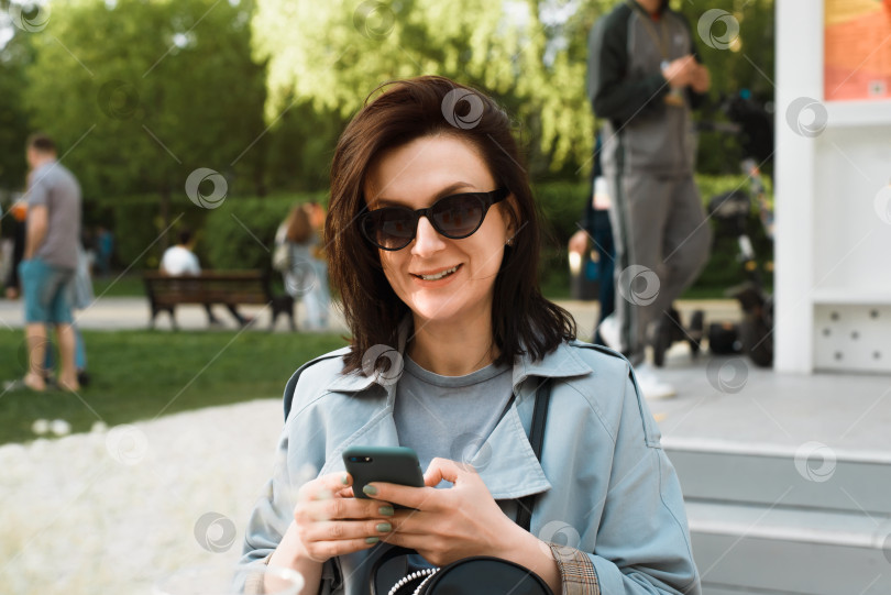 Скачать Болтаем по телефону. Улыбающаяся женщина, пользующаяся мобильным телефоном в уличном кафе на открытом воздухе. Счастливая дама в очках отдыхает в кафе и держит в руках смартфон, глядя в камеру. фотосток Ozero