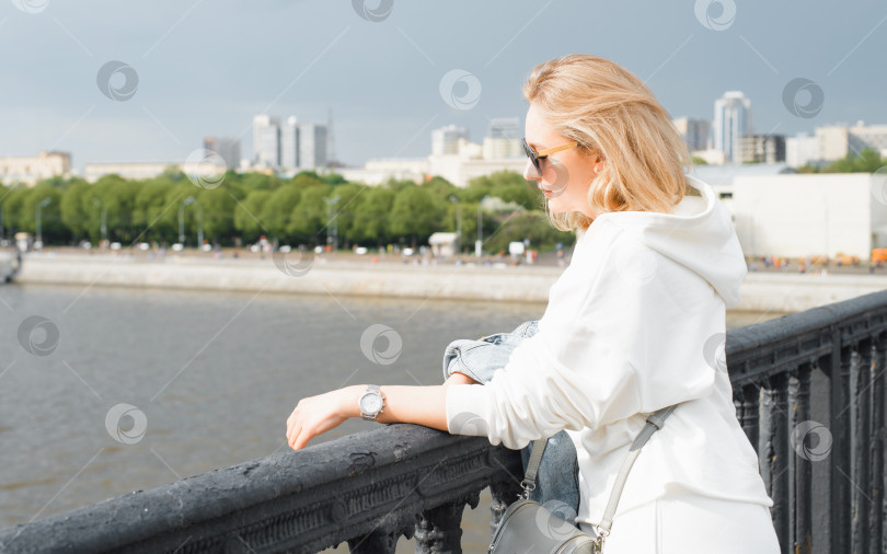 Скачать Портрет женщины в солнцезащитных очках, стоящей на мосту и смотрящей на набережную и реку. Молодая блондинка в повседневной, легкой одежде на прогулке по городу. Путешествуйте по туризму солнечным летним днем. фотосток Ozero