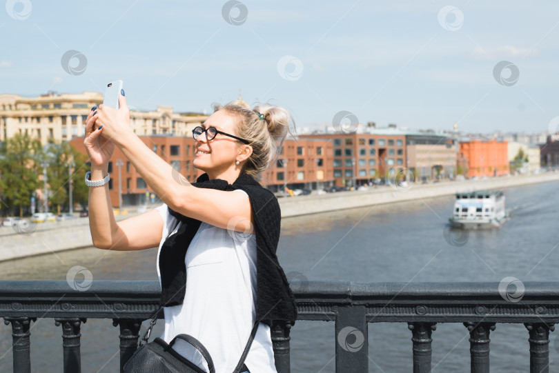 Скачать Счастливая женщина делает селфи на камеру мобильного телефона на открытом воздухе. Улыбающаяся туристка со смартфоном в руках стоит на мосту. Размытый фон реки, городского пейзажа и корабля. Концепция туристической поездки, отпуска фотосток Ozero