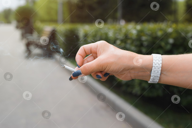 Скачать Женщина курит. Крупным планом женская рука, держащая дымящуюся сигарету на открытом воздухе. Нездоровый образ жизни, вредная привычка. Размытый фон фотосток Ozero